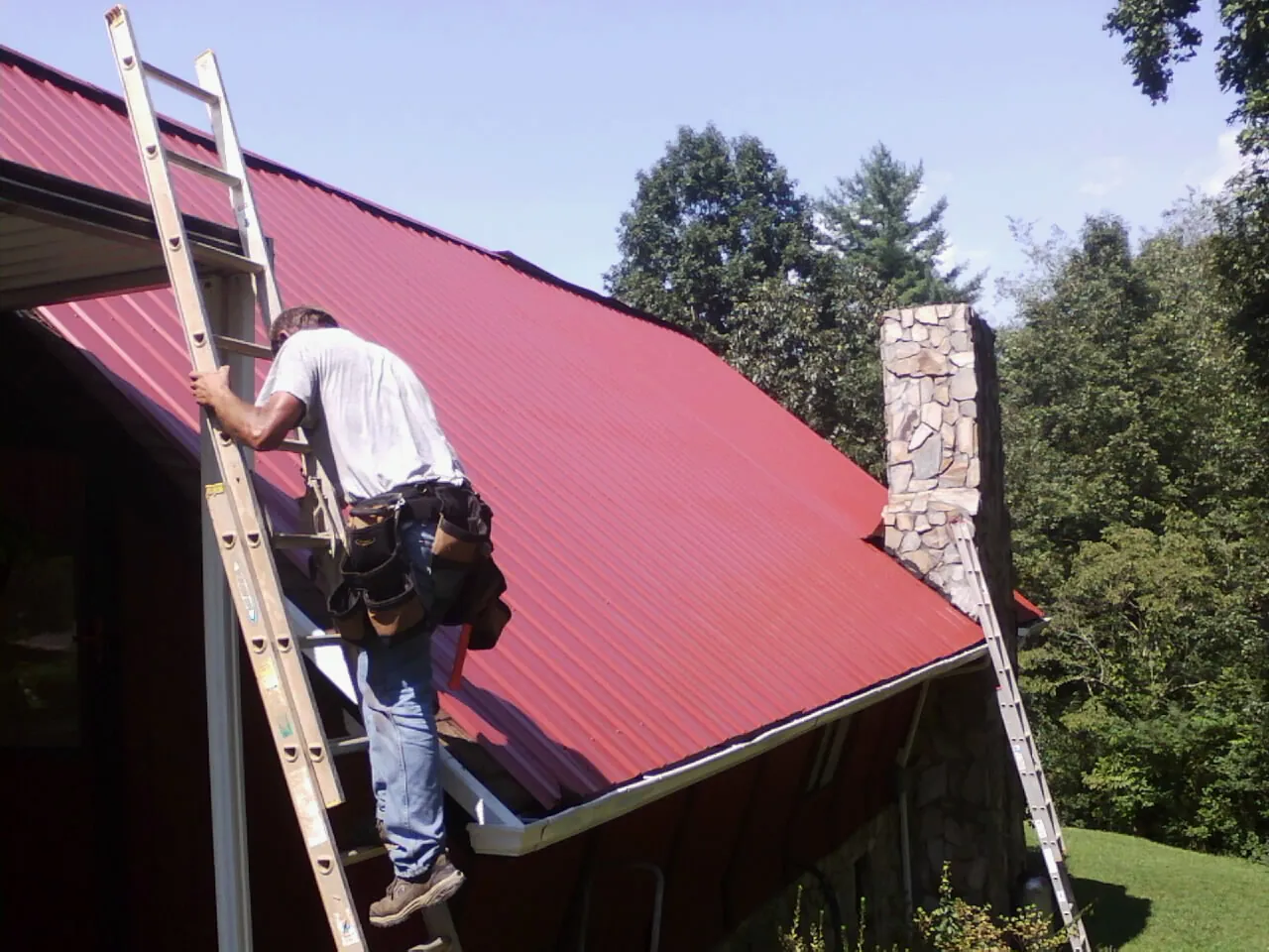 Roofer on a ladder beside a red metal roof and stone chimney — Soffit & Fascia Repair by Roofing Brothers in Seligman